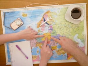 世界地図の上にカメラ、コーヒー、ノートとペンがあり、3人の手がヨーロッパの国を指さしている